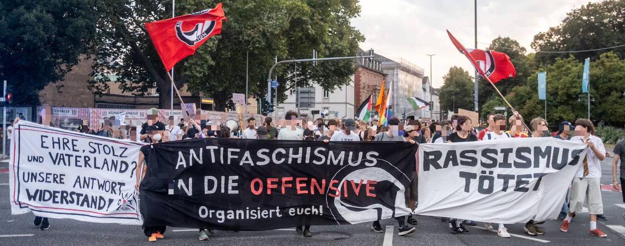 Abgebildet ist eine Demonstration in Gießen. Auf den drei Transparenten steht:
Ehre, Stolz, Vaterland? Unsere Antwort Widerstand!, Antifaschismus in die Offensive Organisiert euch! sowie Rassismus Tötet. Im Hintergrund sind verschiedene Flaggen zu sehen.