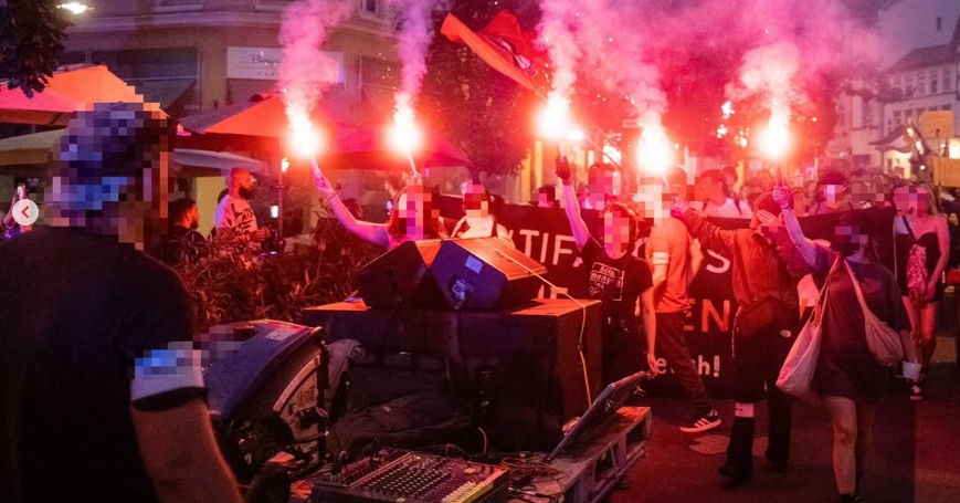 Eine Gruppe von Aktivist*innen hält rot brennende Bengalos über ihren Köpfen. Dahinter sind zahlreiche Demonstrant*innen. Vorne wird ein Transparent gehalten. Im Vordergrund ist ein Technikwagen zu sehen.